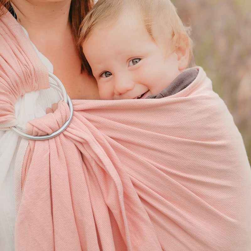 Un bébé porté en sling "rose abélia" il sourit et est confortablement blotti contre sa maman.