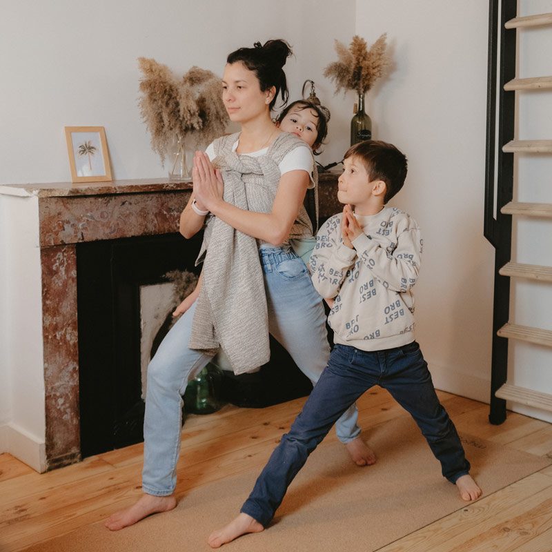 Une femme brune, dans son salon fait une posture de yoga simple avec un bébé en écharpe dans le dos. Un garçon, probablement son fils, la regarde et fait la même posture à coté d' elle.